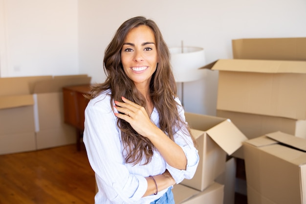 Heureuse belle jeune femme se déplaçant dans le nouvel appartement, debout devant le tas de boîtes en carton ouvertes, regardant la caméra