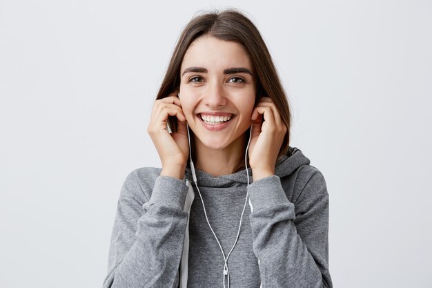 Heureuse belle jeune fille caucasienne aux cheveux noirs avec des cheveux longs en gris à capuche souriant avec des dents, tenant des écouteurs avec les mains, avec une expression de visage excitée et joyeuse