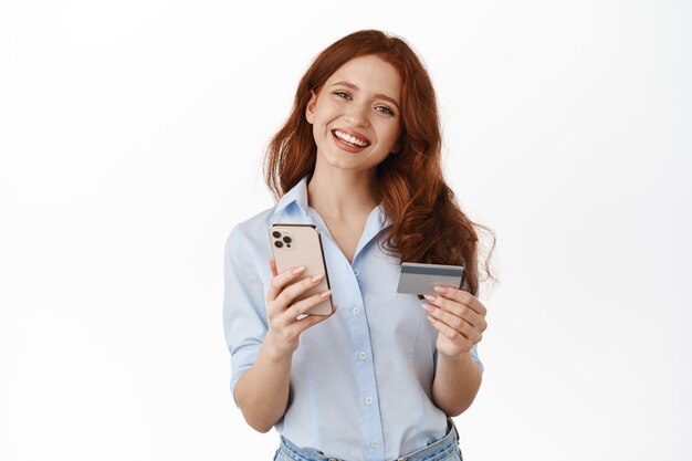 Heureuse commerçante souriante tenant un téléphone portable, une carte de crédit, payant dans l'application pour smartphone, commander quelque chose sur place, vérifiant le compte bancaire, debout sur fond blanc.
