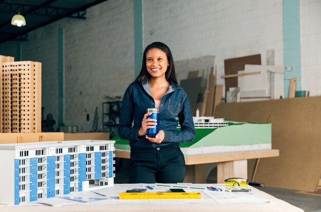 Heureuse dame afro-américaine avec thermos debout près du modèle de bâtiment