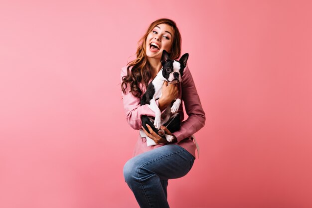 Heureuse dame aux cheveux roux dansant et tenant un chien mignon. Portrait intérieur d'une femme frisée romantique exprimant des émotions positives pendant la séance de portraits avec le bouledogue français.