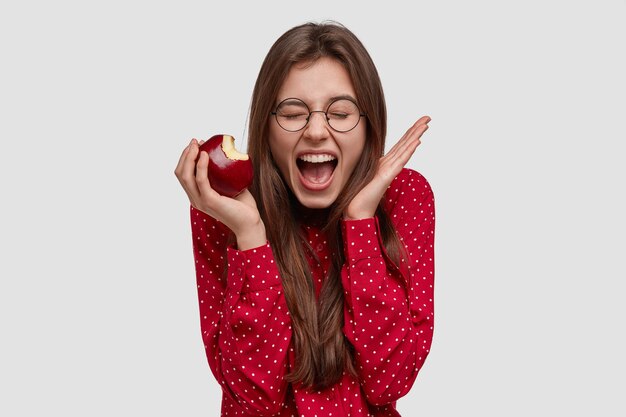 Heureuse dame brune s'exclame et fait des gestes activement, mord une pomme fraîche, garde les yeux fermés, aime une alimentation saine