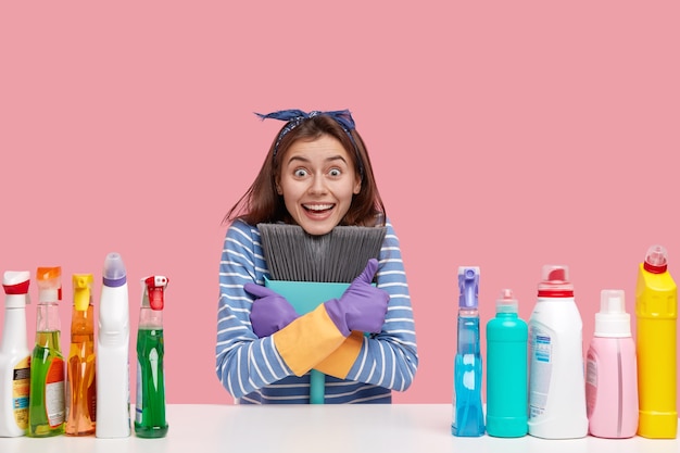 Photo gratuite heureuse dame caucasienne porte un balai près de la poitrine, vêtue de vêtements rayés, regarde avec les yeux pleins de bonheur, a de nombreuses bouteilles de détergent sur la table