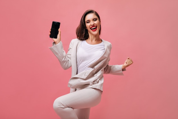 Heureuse dame en costume beige pose avec téléphone sur fond rose. Fille joyeuse en tenue de bureau et avec des lèvres rouges tient le smartphone.