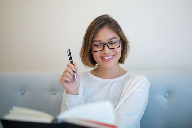 Heureuse dame élevant un stylo et un livre de lecture sur un canapé à la maison