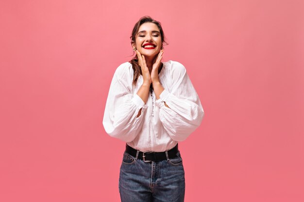 Heureuse dame en jeans et chemise blanche souriant sur fond rose. Enthousiaste belle femme aux cheveux noirs en robe à la mode se penche sur la caméra.