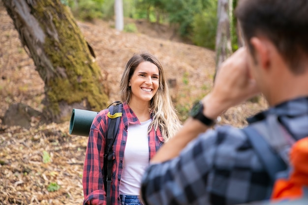 Heureuse dame posant et souriant sur la route en forêt. Homme méconnaissable prenant une photo de sa petite amie. Les touristes marchent ensemble dans les bois et s'amusent. Concept de tourisme, d'aventure et de vacances d'été