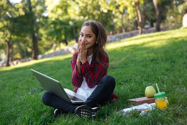 Heureuse écolière brune assise sur l'herbe avec un ordinateur portable