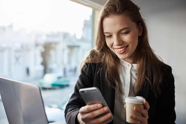 Heureuse élégante étudiante assise dans un café, boire des boissons et messagerie via smartphone, travaillant sur un projet avec un ordinateur portable