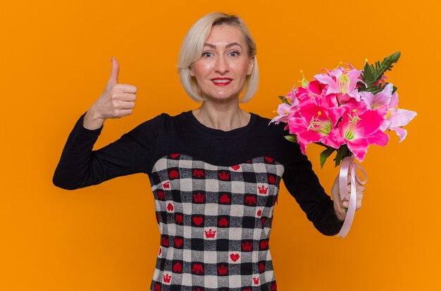 Heureuse et excitée jeune femme en belle robe tenant un bouquet de fleurs à l'avant souriant montrant les pouces vers le haut célébrant la journée internationale de la femme debout sur le mur orange