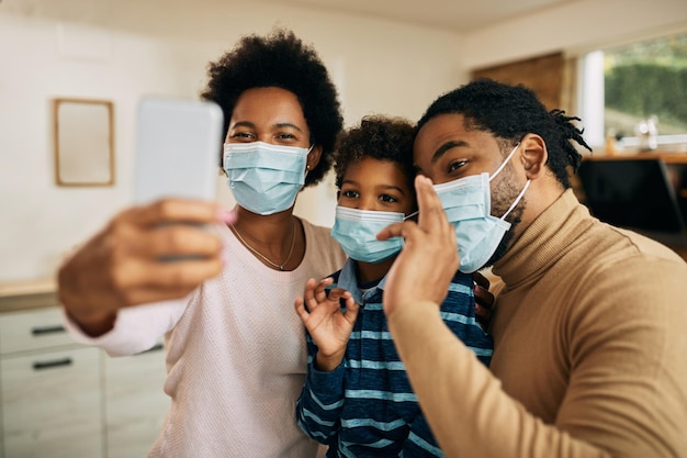 Heureuse famille afro-américaine avec des masques faciaux ayant un appel vidéo sur un smartphone à la maison