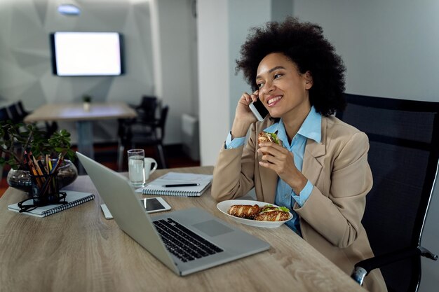 Heureuse femme d'affaires afro-américaine parlant au téléphone tout en mangeant un sandwich au bureau