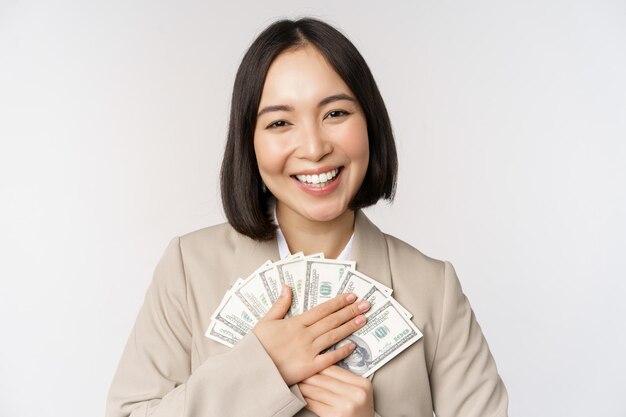 Heureuse femme d'affaires asiatique détenant de l'argent étreignant de l'argent en dollars et souriant debout sur fond blanc en costume