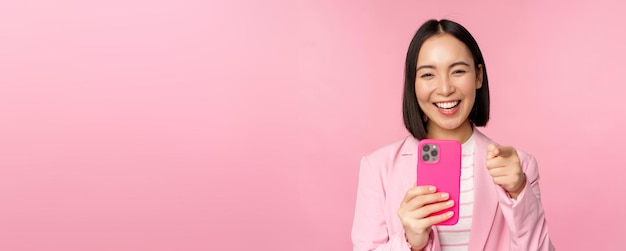 Heureuse femme d'affaires asiatique riant du doigt pointé vers vous et enregistrant une vidéo en prenant des photos sur un smartphone à l'aide d'un fond rose de téléphone portable