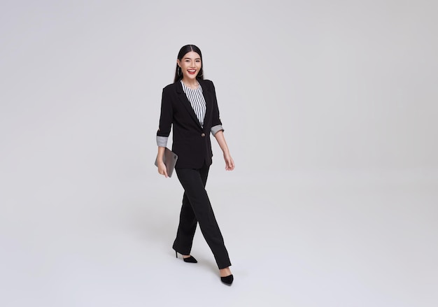 Heureuse femme d'affaires asiatique sourire en costume formel tenant une tablette et marchant sur fond gris