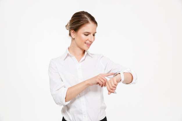 Heureuse femme d'affaires blonde à l'aide de sa montre-bracelet sur mur blanc