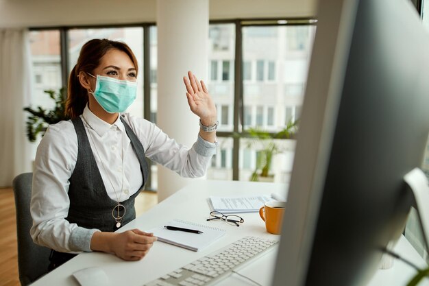 Heureuse femme d'affaires portant un masque facial et agitant tout en ayant une conversation vidéo sur ordinateur au bureau