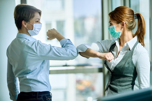 Heureuse femme d'affaires portant des masques faciaux et se cognant le coude lors d'une réunion au bureau pendant l'épidémie de coronavirus