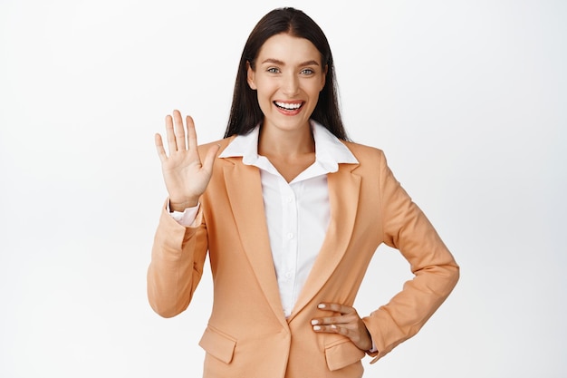 Heureuse femme d'affaires réussie disant bonjour en agitant la main et souriant saluant les clients debout en costume sur fond blanc