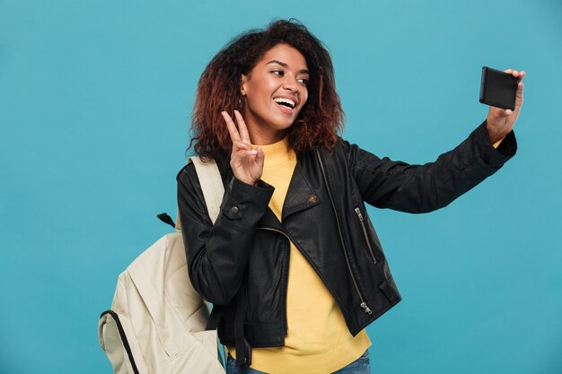 Heureuse femme africaine en veste en cuir avec sac à dos faisant selfie