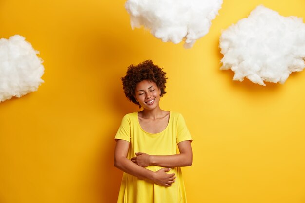 Heureuse femme afro-américaine embrasse le ventre de femme enceinte, exprime son amour pour l'enfant à naître, sourit joyeusement, profite des derniers mois de grossesse, isolé sur un mur jaune. La future mère embrasse le ventre