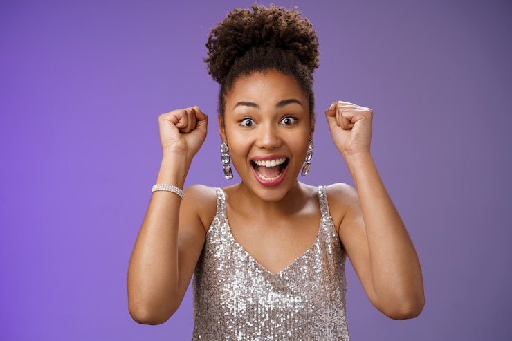 Photo gratuit heureuse femme afro-américaine étonnée en robe argentée à la mode remportant le premier prix célébrant avec joie la levée des poings serrés dansant triomphant joyeusement en s'amusant, debout sur fond bleu.