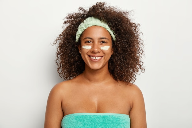 Heureuse femme afro-américaine met la crème pour le visage sous les yeux, a la peau sèche, bénéficie de traitements de rajeunissement, enveloppée dans une serviette, porte un bandeau doux, sourit positivement