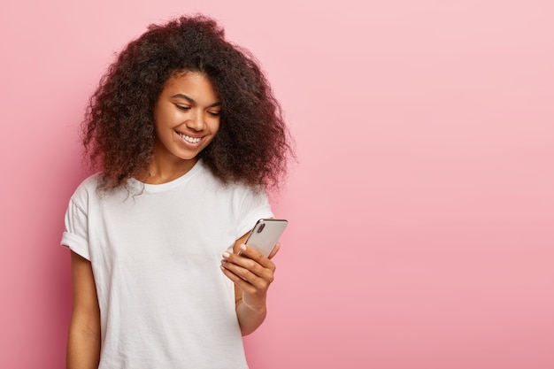 Heureuse femme afro-américaine millénaire avec des boucles sombres, regarde une vidéo drôle sur un téléphone intelligent