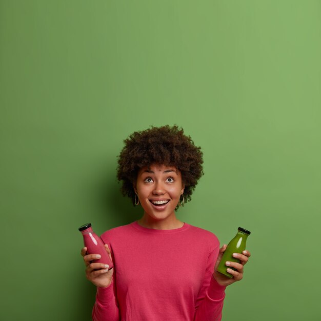 Heureuse femme afro-américaine a un repas sain, détient un smoothie aux fraises et aux épinards dans des bouteilles en verre, boit des boissons de désintoxication, suit un régime, mène un mode de vie sain, concentré au-dessus sur un espace vide