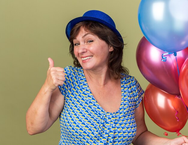 Heureuse femme d'âge moyen en chapeau de fête avec bouquet de ballons colorés souriant montrant les pouces vers le haut célébrant la fête d'anniversaire debout sur un mur vert