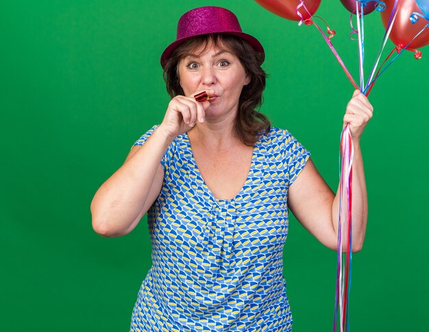 Heureuse femme d'âge moyen en chapeau de fête tenant des ballons colorés soufflant un sifflet heureux et joyeux célébrant la fête d'anniversaire debout sur un mur vert