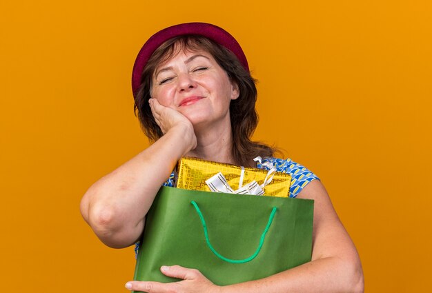 Heureuse femme d'âge moyen en chapeau de fête tenant un sac en papier avec des cadeaux d'anniversaire avec les yeux fermés souriant joyeusement célébrant la fête d'anniversaire debout sur un mur orange