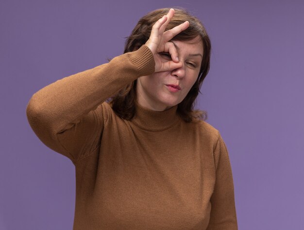 Heureuse femme d'âge moyen en col roulé marron faisant signe ok regardant à travers ce signe un clin de œil debout sur le mur violet