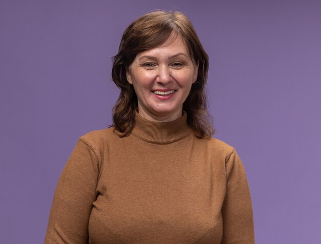 Heureuse femme d'âge moyen en col roulé marron souriant largement debout sur le mur violet