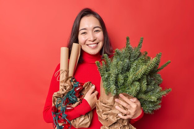 Heureuse femme asiatique brune a une humeur festive porte des branches d'épinette en papier roulé et une guirlande rétro sourit agréablement porte des poses de pull décontractées sur fond rouge vif Décoration de vacances
