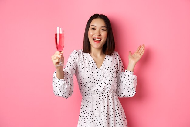 Heureuse femme asiatique célébrant en disant un toast à la fête levant une coupe de champagne et souriante debout ...
