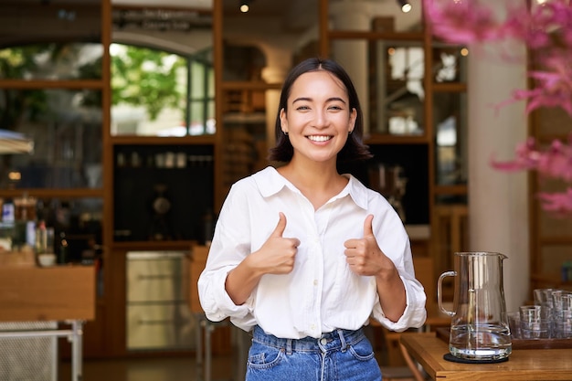 Heureuse Femme Asiatique Montre Le Pouce En L'air Approuve Aime Qch Bon Se Tient Près De L'entrée Du Café Et Lui Donne Re