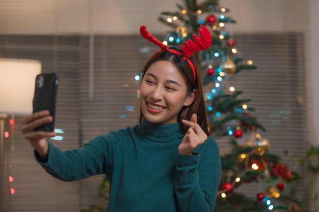Heureuse femme asiatique souriante appel vidéo via smartphone pour célébrer avec ses amis ou sa famille pendant noël à la maison en faisant un mini geste cardiaque
