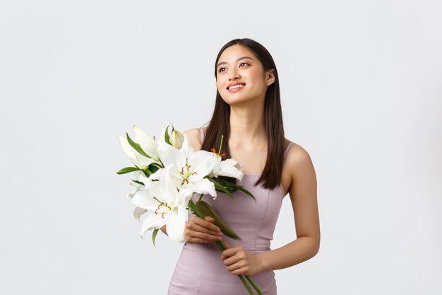 Heureuse femme asiatique souriante en robe élégante, regardant dans le coin supérieur gauche et tenant le bouquet de lys