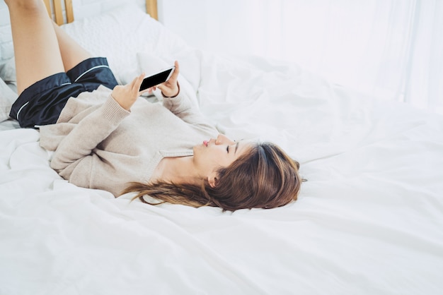 Heureuse femme asiatique utilise un téléphone intelligent avec un écran vide vide noir sur le lit le matin