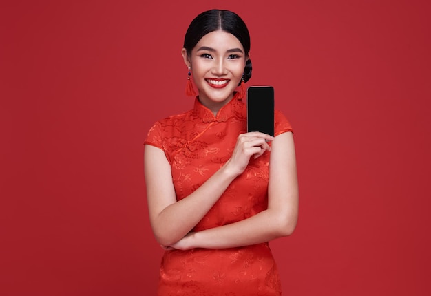 Heureuse femme asiatique vêtue d'une robe traditionnelle cheongsam qipao montrant un téléphone portable isolé sur fond rouge.