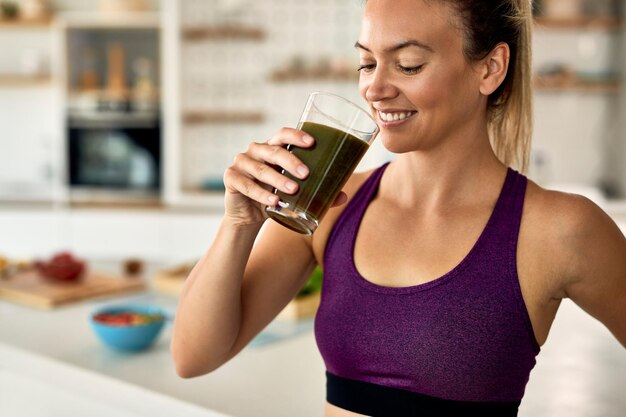 Heureuse femme athlétique buvant un smoothie vert dans la cuisine