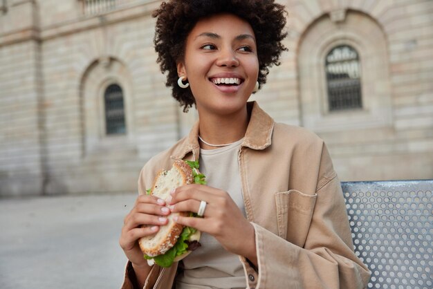 Heureuse femme aux cheveux bouclés est assise à l'extérieur mange un délicieux sandwich regarde joyeusement loin préfère la malbouffe vêtue de vêtements décontractés a une pause déjeuner après une promenade dans la ville Les gens mode de vie nutrition