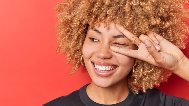 Heureuse femme aux cheveux bouclés heureuse fait un clin d'œil fait un geste de paix sourit largement regarde ailleurs a une expression joyeuse concentrée loin isolé sur fond rouge espace vide pour votre contenu publicitaire