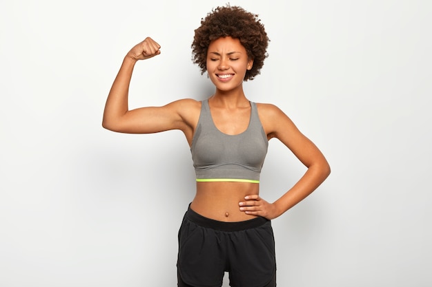 Heureuse femme aux cheveux bouclés lève le bras, montre les biceps, démontre les muscles, a une silhouette mince, porte un haut et un short décontracté, des modèles sur un mur blanc