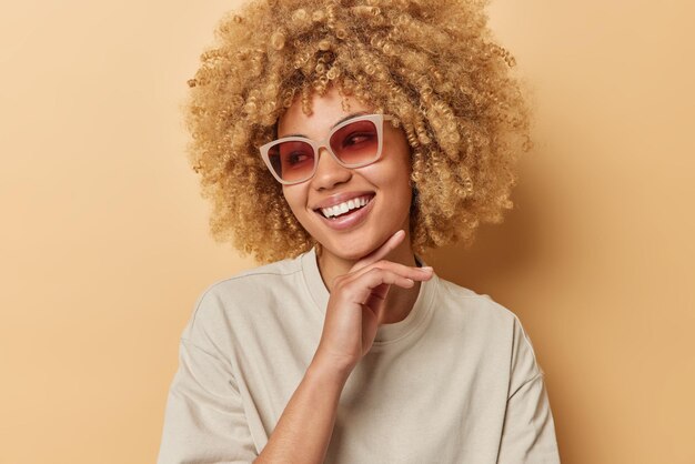 Heureuse femme aux cheveux bouclés touche la mâchoire sourit doucement regarde loin porte des lunettes de soleil et un t-shirt décontracté se sent heureux habillé en t-shirt décontracté isolé sur fond marron Concept de joie des gens