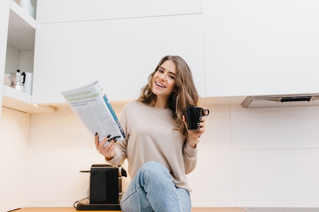 Heureuse femme aux cheveux longs avec une expression de visage heureux, lisant des nouvelles le matin