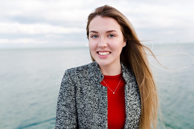 Heureuse femme aux cheveux longs souriante avec de grands yeux bleus en chemise rouge et manteau gris se promène près de la mer