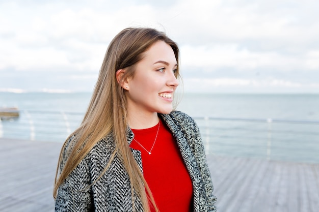 Heureuse femme aux cheveux longs souriante avec de grands yeux bleus en chemise rouge et manteau gris se promène près de la mer