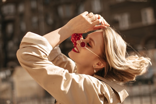 Heureuse femme blonde cool joue les cheveux à l'extérieur Dame excitée avec des lèvres rouges en trench-coat beige sourit à l'extérieur